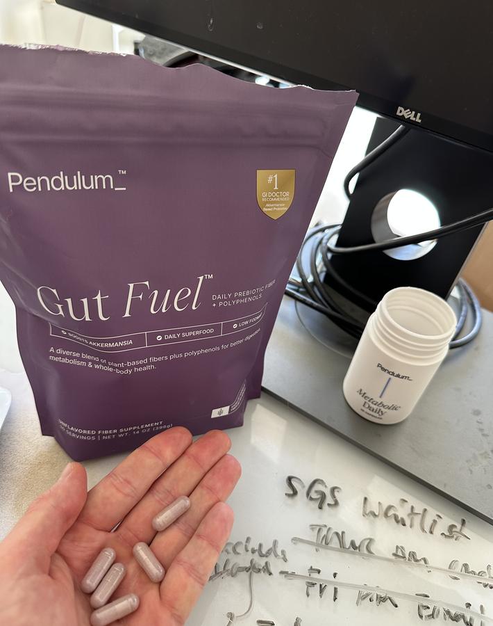 Metabolic Daily capsules held in a palm at a desk, with the open jar and a Gut Fuel pouch behind